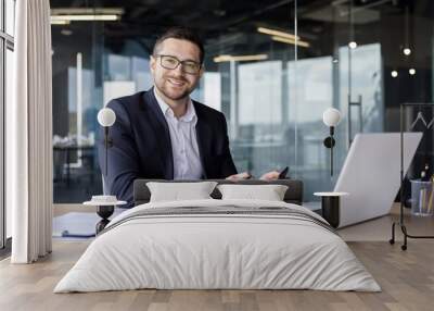 Portrait of a young businessman, accountant, financial analyst working in the office with a calculator and documents, sitting at a desk with a laptop and smiling at the camera Wall mural