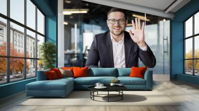 Portrait of a young businessman in a suit who sits in the office in front of the camera, communicates online, greets with his hand Wall mural