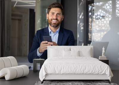 Portrait of a young businessman sitting in a suit at a desk inside the office, holding a mobile phone, smiling and looking at the camera Wall mural