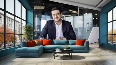 Portrait of a young businessman smiling at the camera, sitting at a desk in the office and working with documents and an invoice Wall mural