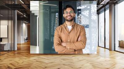 Portrait of a young Indian man wearing glasses and a brown shirt standing in a modern office, crossing his arms over his chest and looking confidently at the camera Wall mural