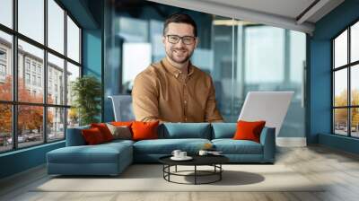 Portrait of a young male office worker, businessman smiling and posing at the camera and working on a laptop while sitting at a desk Wall mural