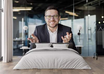 Portrait of a young male teacher teaching online via video call from the office. Sits in front of the camera and explains with a smile, gesturing with his hands Wall mural