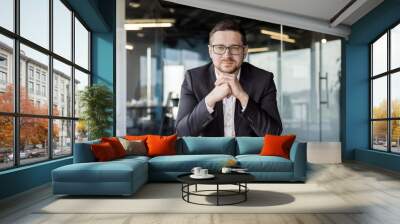 Portrait of confident young man businessman, executive director, boss sitting in office at table in business suit and looking at camera with folded hands together Wall mural