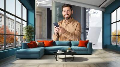 Portrait of mature successful businessman inside office, male looking at camera smiling, worker in shirt holding phone, using online application Wall mural