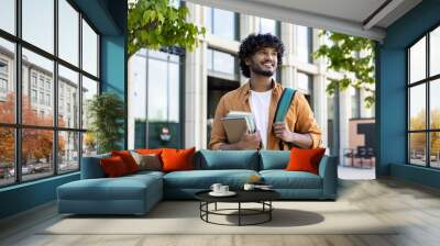 Portrait of young Indian male student, applicant standing outside campus, holding books and backpack, preparing for exam. Smiling and confidently looking to the side Wall mural