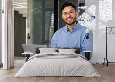 Portrait of young successful businessman inside office, satisfied joyful Indian man looking at camera, worker at workplace using laptop, typing on keyboard, financier accountant in shirt Wall mural