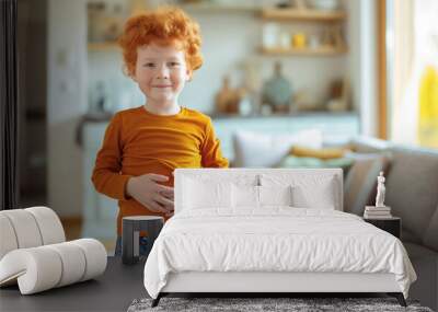 Red-haired boy in orange shirt holding his stomach, standing in a bright, cozy living room, concept of stomach ache, parasites, and digestive issues in children Wall mural