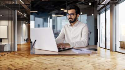 Serious young Indian businessman man working focused on laptop while sitting in office at desk Wall mural