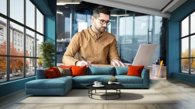 Serious young man accountant, auditor, analyst works in the office at the table behind the laptop and with documents. Writes invoices, makes analysis, fills out financial reporting and taxation Wall mural