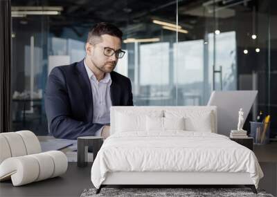 Serious young man accountant, financier and economist working in office at laptop and with documents, holding calculator in hands, concentrating on accounts Wall mural