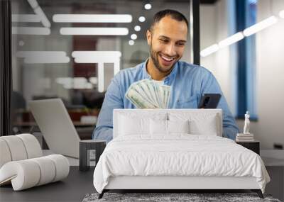 Smiling hispanic young man sitting in a formal office at a table, holding cash money banknotes in his hands, using a mobile phone Wall mural