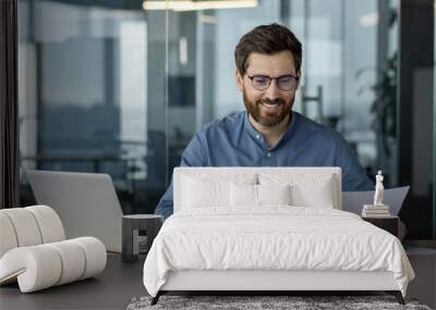 Smiling young man businessman in glasses works in office with laptop and documents, studies data, conducts analysis and examination Wall mural