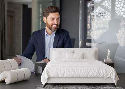 Smiling young man working in office with documents, sitting at desktop with laptop and holding online meeting with clients and partners, signing agreement and invoices Wall mural