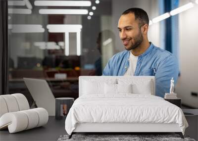 Smiling young Muslim man businessman, designer, freelancer, programmer in blue shirt working in office with laptop, sitting at office table and typing on keyboard Wall mural