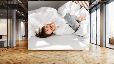 Cute little happy child boy playing in parents bed having fun at home at sunny morning. Light and shadows Wall mural