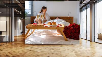 Romantic breakfast in bed on Valentine Day.  Couple having fun in bed celebrating holiday with champagne, gift, croissants and bouquet of red roses. Wall mural