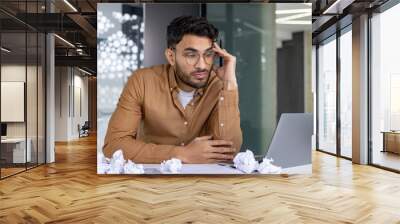 Stressed businessman working at desk with crumpled papers and laptop in office Wall mural