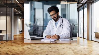 Successful smiling doctor working inside modern clinic office, hindu man in white medical coat working in medical office sitting at table with laptop Wall mural