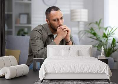 Upset man sitting alone at home, depressed hispanic man on sofa in living room in casual clothes thinking about difficult decision Wall mural