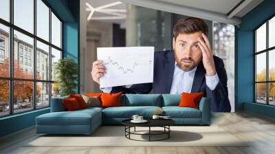 Worried and upset young man in a suit sitting at an office desk in front of the camera and showing documents and graphs while holding his head in frustration Wall mural