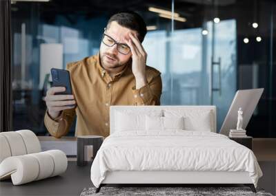 Worried young male businessman received bad news on phone, sitting in office at desk and upset reading message, mail, holding head, thinking about problem solution Wall mural