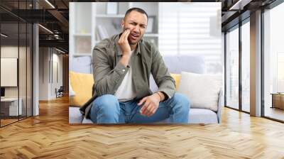 Young hispanic man suffering from toothache sitting on sofa at home and holding hand to cheek Wall mural