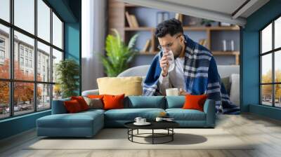 Young man sitting on sofa covered with blanket, drinking hot beverage and blowing nose, feeling unwell and cold Wall mural