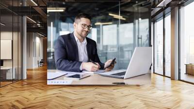 Young man working in the office on paper work, holding a calculator and processing data, bills and documents online via laptop Wall mural