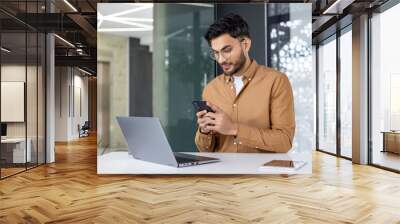 Young professional checking phone while working on laptop in modern office Wall mural