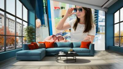 UV protective eyewear concept. Cheerful millennial woman in white shirt choosing new fashionable and functional sunglasses. Female customer shopping comfortable pair of sunnies at local optical store Wall mural