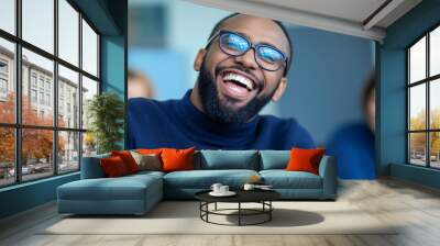 A cheerful businessman in a navy blue sweater and glasses laughs joyfully in an office setting. His expression radiates positivity and happiness, capturing a candid moment of workplace camaraderie. Wall mural