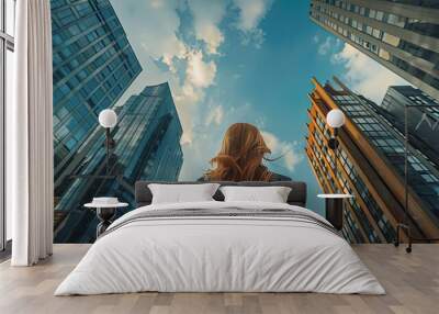 A woman gazes up at towering skyscrapers against a bright blue sky, embodying ambition and urban exploration in a modern city. Wall mural