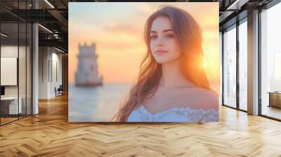 A serene portrait of a woman in a white dress by the sea, illuminated by a golden sunset, with a historic tower in the background. Wall mural