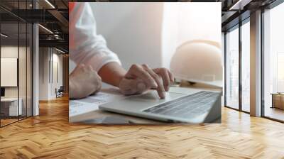 A person is typing on a laptop computer with a construction helmet on the desk. The scene suggests that the person is working on a construction project or related task Wall mural