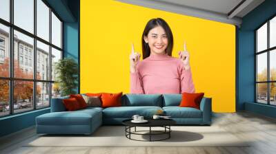 Asian smiling young woman finger Pointing up for advertise, banner or showing on isolated yellow color background, wearing winter cozy sweater indoors studio, happiness and fun, copy space concept Wall mural