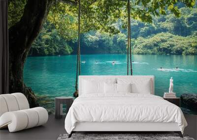 A wooden swing hanging from the tree beside an emerald blue lake, surrounded by lush greenery and swimmers in the clear water. The swing is made of natural wood with intricate rope design  Wall mural