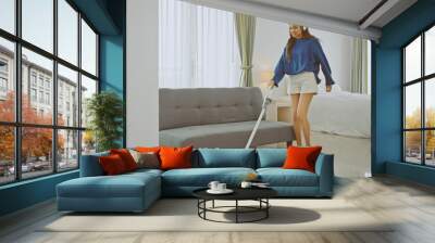 Young woman holding vacuum cleaner, singing and cleaning in her bedroom. Wall mural