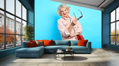 cheerful happy blonde old lady holding sunglasses and having a rest in the studio with blue wall. close up portrait. isolated blue background. people and lifestyle concept Wall mural