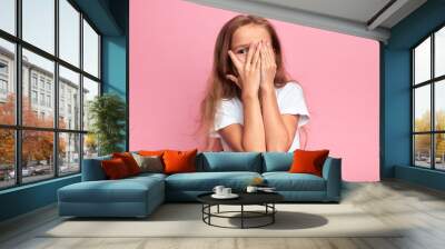 emotional little girl covers her face with her hand isolated over pink background, child watching horror film, movie, reaction, facial expression. isolated pink background Wall mural