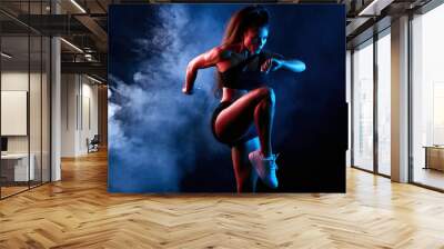 Young serious confident attractive sporty girl runner in shorts and top keeps her leg raised while warming up and stretching indoors. isolated black background, lifestyle Wall mural