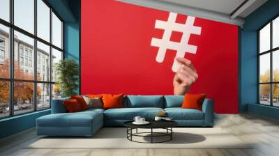 A person holding a large hashtag symbol against a vibrant red background, symbolizing social media trends and connectivity. Wall mural