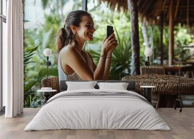 Side view of smiling female digital nomad sitting on wicker high stool and taking self portrait on cellphone in cafe at tropical resort while on summer trip to Bali : Generative AI Wall mural