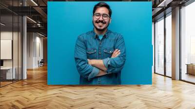 Young hispanic man wearing blue shirt and glasses looking at camera with positive confident smile holding arms crossed isolated on blue background : Generative AI Wall mural