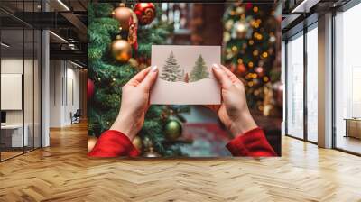 First person view photo of hands holding a christmas greeting card in front of a xmas tree.. Wish you a merry christmas postcard. Wall mural