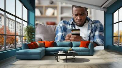 Worried African American man looking at mobile phone. Sitting on the couch upset and holding a credit card, Generative AI Wall mural