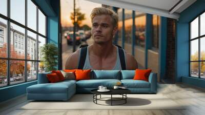 A handsome muscular blond man walking along the city street during sunset, showcasing his fitness in casual attire. Wall mural