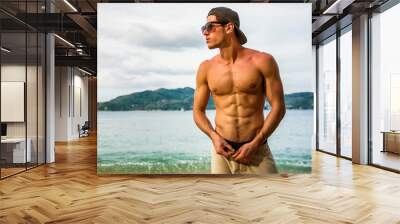 Attractive shirtless athletic young man on beach by the sea, with baseball cap and sunglasses, looking away Wall mural