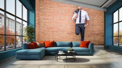 Portrait of stylish young man wearing business suit, standing in modern city setting, leaning against brick wall Wall mural