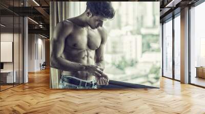 Sexy handsome young man standing shirtless in his bedroom next to window curtains, adjusting wrist watch Wall mural